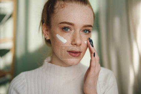 Photo for Close up beauty girl with freckles applying moisturizing skincare cream, lotion or mask for skin lifting and anti-aging detoxifying effect. Looking at camera - Royalty Free Image