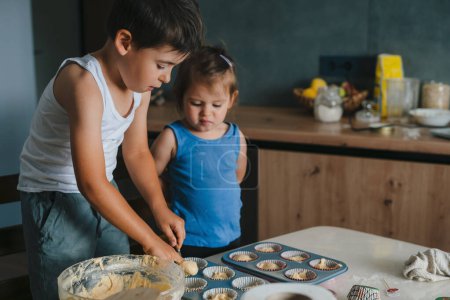 Photo for Little boy and girl pouring batter into cupcake molds while standing in the kitchen. Homemade cooking, food - Royalty Free Image
