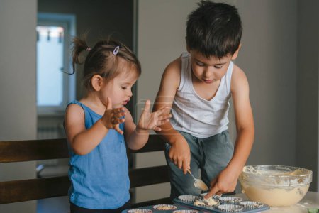 Photo for Focused boy pouring raw batter into metal cupcake while standing next to his sister in light kitchen at home. The process of preparation for baking. - Royalty Free Image