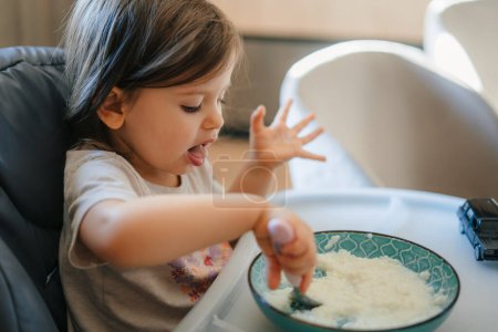 Happy little child enjoying healthy breakfast, eating porridge sitting in high chair. Child development concept