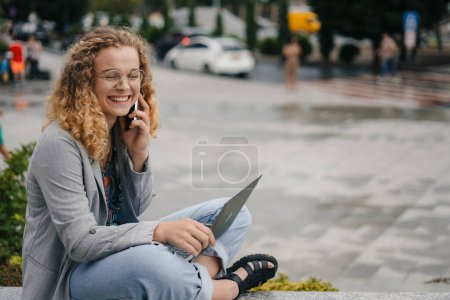 Portrait of happy young woman working remotely online sitting with laptop outside in same time speaking with smile on phone with her superior