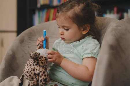 Porträt eines kleinen kaukasischen Mädchens mit blauem Filzstift auf dem heimischen Sofa, das ein Stofftier beschmutzt. Kindheit Freizeit Spaß spielen Lernentwicklung
