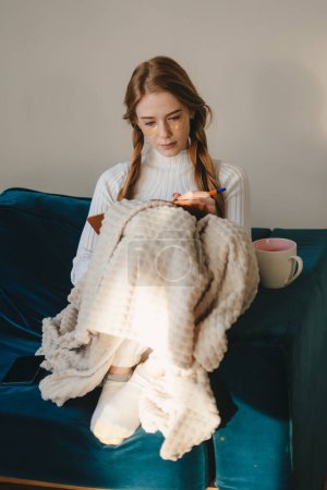 Femme rousse réfléchissant et écrivant des notes sur un cahier, assise sur un canapé confortable et recouverte d'une couverture dans son salon