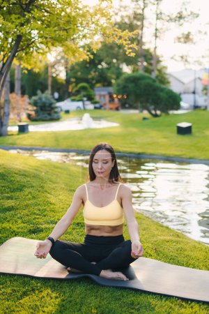 Jeune femme en forme adulte pratiquant le yoga sur tapis de gymnastique dans le parc vert sur l'herbe. Faire des exercices de respiration matinale assis jambes croisées en vêtements de sport.