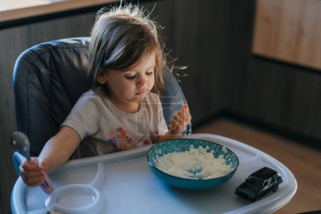 Petite fille adorable affamée qui mange du porridge dans un bol au petit déjeuner. Dégustation de riz sucré délicieux au lait, repas équilibré pour enfants concept.
