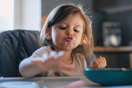 Happy small cute kid girl eating morning meal from bowl feeling hungry sitting at high chair. Balanced meal for children concept.