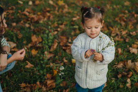 Cute toddler girl among autumn park picking wild flowers in park or forest on a spring day. Little kid exploring nature. Outdoor activities for children