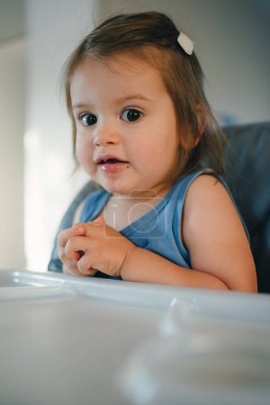 Small cute little toddler brunette caucasian girl sitting in baby chair looking at camera. Self-feeding concept