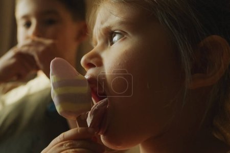 Ein kleines Mädchen isst freudig einen Eibisch und streckt die Zunge heraus. Sie scheint glücklich und verspielt, genießt einen lustigen Moment des Teilens und der Freude