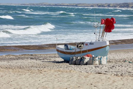 Téléchargez les photos : A fishing boat on the beach with red flags in it. The sea is visible in the back. The sun I shining and the sky and sea is blue. - en image libre de droit