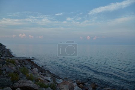 Photo for Landscape of sean and beach at dusk. The sky is blue with small white clouds. The the foreground there is big rocks. - Royalty Free Image