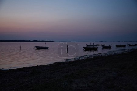 Photo for Dinghies on the water at dusk photographed in perspektive. In the foreground there is a  slice og of beach.In the background there is land. The sky and water is pastel colored in blue and rosa. Vertikal. - Royalty Free Image