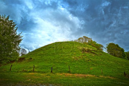 Photo for A green burial mound against a dramatic sky. Both in front and back there is green trees. - Royalty Free Image