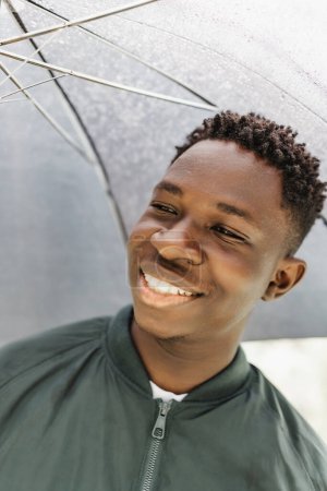 Young African American man with umbrella, under rain, smiling on street outdoor. Fall or spring season
