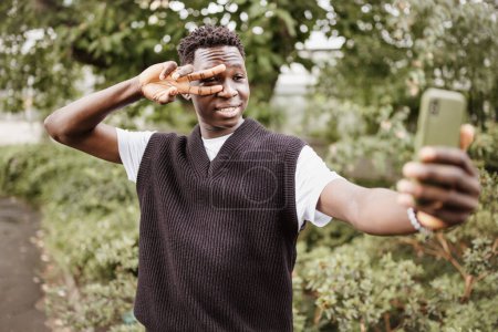 Photo for Young happy African American man taking selfie on his mobile phone, show v sign near his eye, smiling in park or street outdoor. Fall or spring season - Royalty Free Image
