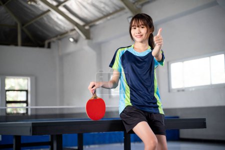 Photo for Female ping pong player holding red ping pong paddle with thumbs up in indoor building - Royalty Free Image