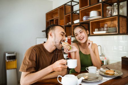 Foto de Feliz asiático pareja bebiendo café en tazas juntos mientras alimentación cada otro desayuno en comedor fondo - Imagen libre de derechos