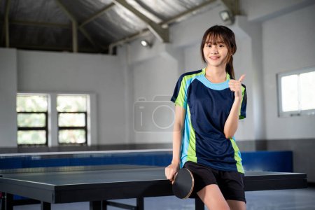 Photo for Female ping pong player holding paddle with thumbs up in indoor building - Royalty Free Image
