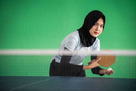 Photo for Young asian girl in hijab serving while playing ping pong on green background - Royalty Free Image