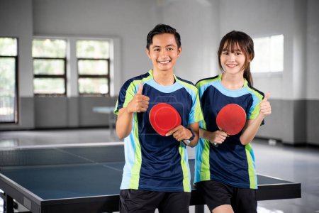 Photo for Two smiling asian athletes holding paddle with thumbs up in front of ping pong table - Royalty Free Image