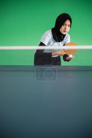 Photo for Asian female athlete in hijab serving while playing table tennis on green background - Royalty Free Image