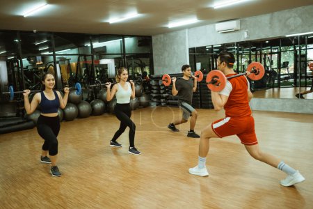 Téléchargez les photos : Male instructor gives an example of back squats with a barbell in front with a young sporty group in the fitness center - en image libre de droit
