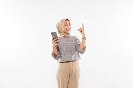 Téléchargez les photos : A young woman with hijab standing raising her hand and holding the phone at her right hand on the white background - en image libre de droit