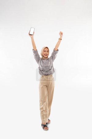 Téléchargez les photos : A moslem woman in shirt and long pants standing raising her arms while holding the phone at her right hand and clenched her left hand on the white background - en image libre de droit