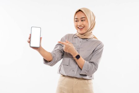 Téléchargez les photos : A beautiful woman with hijab standing with smile and pointing on the phone at her hand on the white background - en image libre de droit