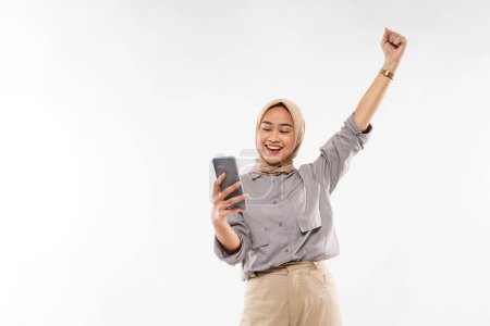 Téléchargez les photos : A young woman with hijab smiling while standing with raised and clenched arm and looking at her phone on the white background - en image libre de droit
