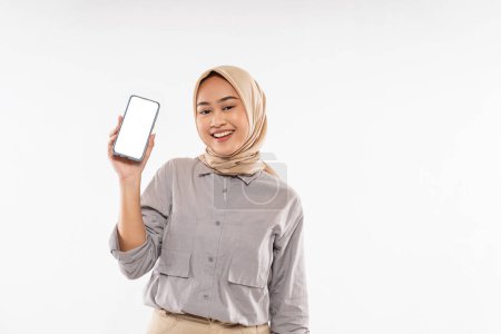 Téléchargez les photos : A beautiful woman with hijab standing with smile while raising and showing the phone at her hand with the white background - en image libre de droit