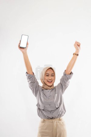 Téléchargez les photos : A young woman with hijab standing raising her arms while holding the phone at her right hand and clenched her left hand on the white background - en image libre de droit
