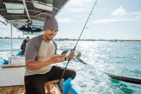 Photo for Fisherman using a smartphone to send messages while fishing at sea on a small fishing boat - Royalty Free Image
