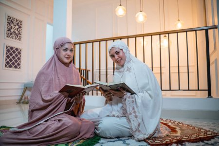 Photo for Two attractive girls wearing Muslim clothes with hijab reciting the Quran sitting on a prayer mat in a room - Royalty Free Image