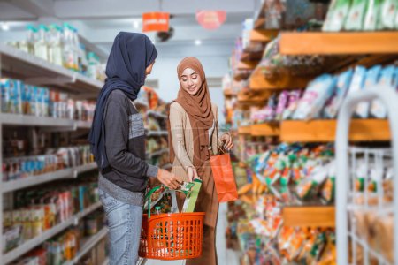 Dos chicas musulmanas poniendo cosas en una canasta mientras compran juntas en un supermercado