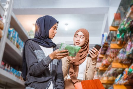 Téléchargez les photos : Deux filles musulmanes discutent tout en vérifiant les marchandises avec des smartphones dans un supermarché - en image libre de droit