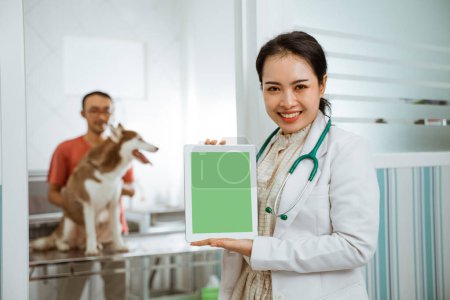 a beautiful vet lean against the door showing the tablet on her hand with the brown siberian dog and its owner at the background