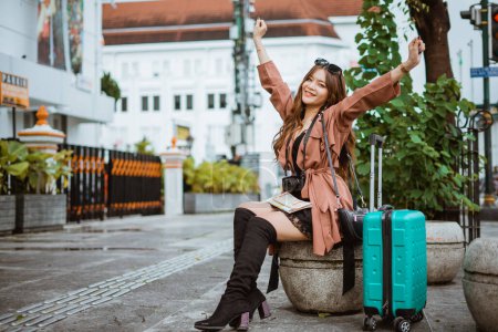 Photo for Female traveller with long brown hair raising her hand and smiling while sitting on the stone seat with the buildings at the background - Royalty Free Image