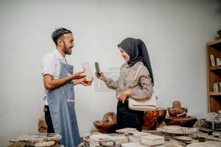 Téléchargez les photos : Serveur masculin sert les clients musulmans féminins tout en scannant quelque chose pour le paiement non monétaire dans un magasin d'artisanat en pierre - en image libre de droit