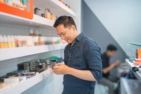 Téléchargez les photos : Mâle vape vendeur regardant son téléphone tout en se tenant à côté de l'étagère du magasin - en image libre de droit