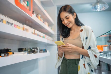 Photo for Beautiful asian shopkeeper checking her phone while standing beside the liquid shelf inside the vape store - Royalty Free Image