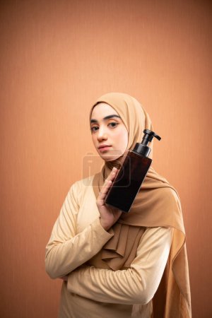Téléchargez les photos : Femme musulmane portant hijab et t-shirt crème à la fois main tenant bouteille de soins de la peau souriant à la caméra en fond orange - en image libre de droit