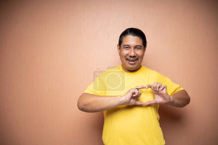 Photo for Old asian man wearing yello tshirt making heart sign with his finger over his chest in plain background isolated - Royalty Free Image