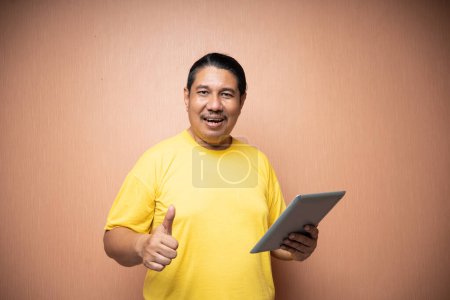 Photo for Old asian man holding tablet with left hand and showing thumb up gesture in plain background isolated - Royalty Free Image