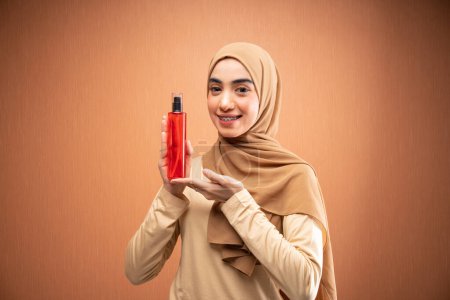 Téléchargez les photos : Femme musulmane portant hijab et t-shirt crème à la fois main tenant bouteille de soins de la peau souriant à la caméra en fond orange - en image libre de droit