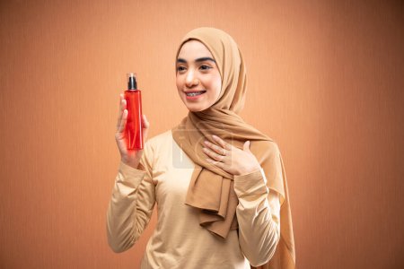 Téléchargez les photos : Femme musulmane portant hijab et crème t-shirt tient bouteille de soins de la peau souriant latéralement dans fond orange et la main gauche dans sa poitrine - en image libre de droit