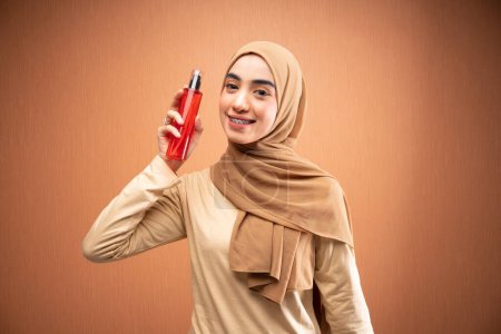 Téléchargez les photos : Femme musulmane portant hijab et t-shirt crème tenant bouteille de soins de la peau souriant à la caméra en fond orange - en image libre de droit
