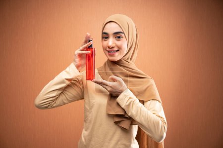 Téléchargez les photos : Femme musulmane portant hijab et t-shirt crème tenant bouteille de soins de la peau souriant à la caméra en fond orange - en image libre de droit