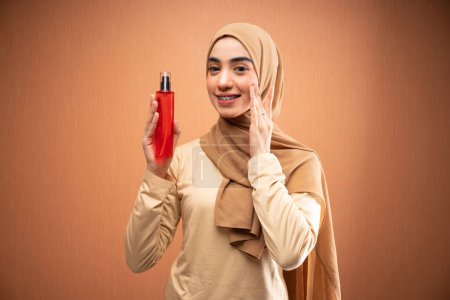 Téléchargez les photos : Femme musulmane portant hijab crème et t-shirt souriant tout en appliquant des soins sur son visage et tenant bouteille souriant à la caméra en fond orange - en image libre de droit