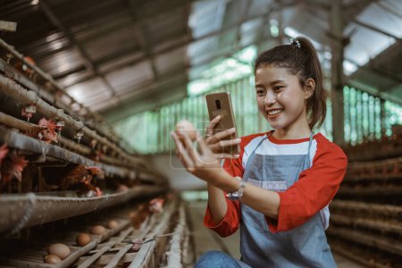 Téléchargez les photos : Une agricultrice de volaille prend une photo d'œufs avec téléphone portable dans un poulailler - en image libre de droit